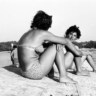 Here's a brief summary of the image: Two women sit on a stone ledge by the water.
