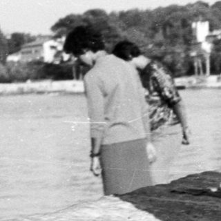 Two women in swimsuits sit on a stone ledge by the sea.