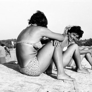 Two women in swimsuits sit on a stone ledge by the sea.