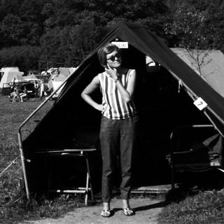 Woman stands near tents in a grassy, wooded area.