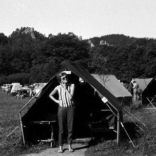 Woman stands near tents in a grassy, wooded area.