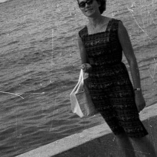 Woman stands by water with boat and coastline visible.