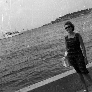 Woman stands by water with boat and coastline visible.