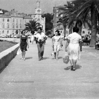 People walk along a waterfront promenade with buildings and palms.