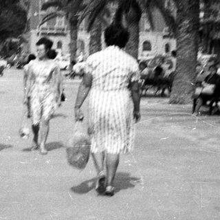 People walk along a waterfront promenade with buildings and palms.