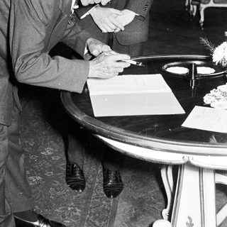 Elderly man signs document, assisted by a younger man.