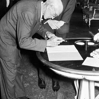 Elderly man signs document, assisted by a younger man.