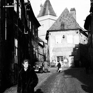 Here's a brief summary of the image: Woman walking down a cobblestone street, vintage cars visible.