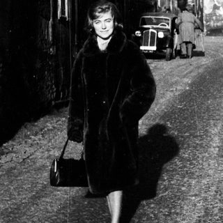 Here's a brief summary of the image: Woman walking down a cobblestone street, vintage cars visible.