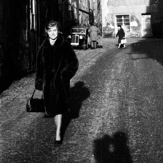 Here's a brief summary of the image: Woman walking down a cobblestone street, vintage cars visible.