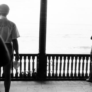 Two figures stand on a balcony overlooking the sea.