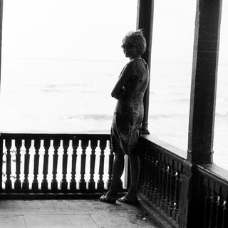Two figures stand on a balcony overlooking the sea.