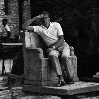 Man seated outdoors, stone wall and table visible.