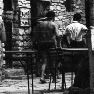 Woman sits in stone chair, people nearby, stone building.