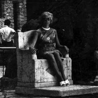 Woman sits in stone chair, people nearby, stone building.