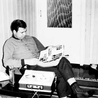 Man reading magazine while seated near vintage stereo equipment.