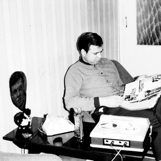 Man reading magazine while seated near vintage stereo equipment.
