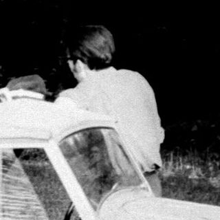 Vintage car and people in a grassy, wooded area.