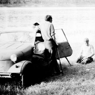 Men relaxing by a car and motorcycle near a lake.