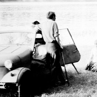 Men relaxing by a car and motorcycle near a lake.