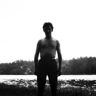 Man stands shirtless by a lake, facing the camera.