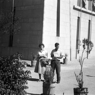 Two people holding potted plants outside a building.