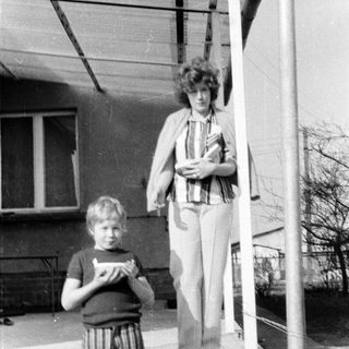 Two people standing outdoors near a building. Woman holding a bag, boy holding a booklet.