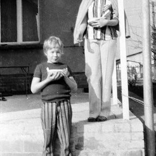 Two people standing outdoors near a building. Woman holding a bag, boy holding a booklet.