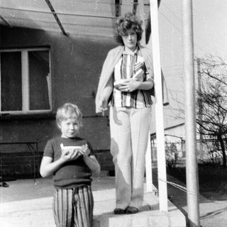 Two people standing outdoors near a building. Woman holding a bag, boy holding a booklet.