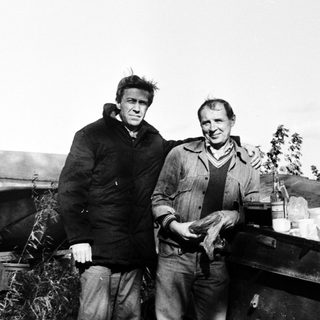 Two men standing outdoors near fishing boats.