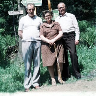 Three elderly people posing outdoors near forest signs.