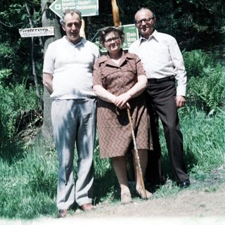 Three elderly people posing outdoors near forest signs.