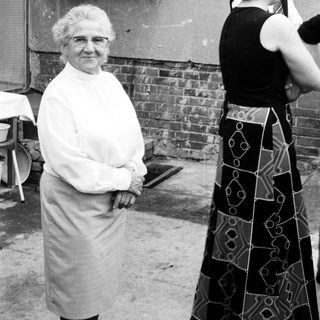 Elderly woman in apron posing outdoors with younger woman taking photo.