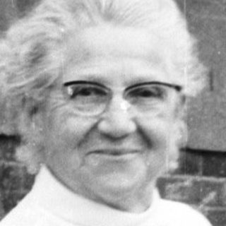 Elderly woman in apron posing outdoors with younger woman taking photo.