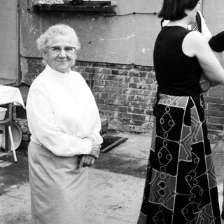 Elderly woman in apron posing outdoors with younger woman taking photo.