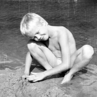 Shirtless person kneeling, drawing in wet sand.