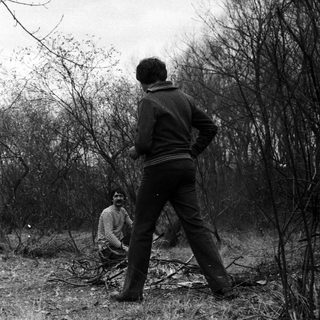 Two men in rural setting, one shirtless, another standing.