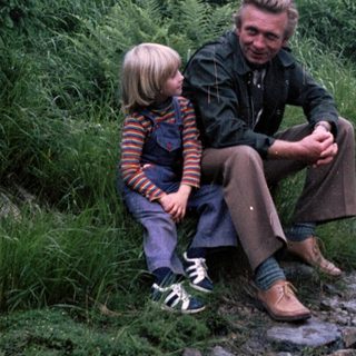 A man and child sitting together on a rocky trail in a forested area.