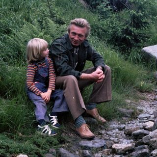 A man and child sitting together on a rocky trail in a forested area.