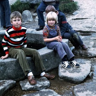Three children sitting on rocks outdoors with adults in background.