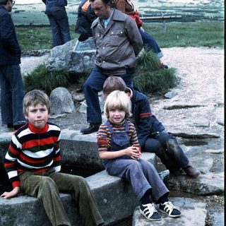 Three children sitting on rocks outdoors with adults in background.
