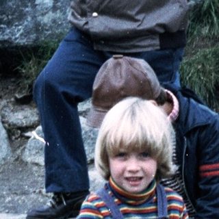 Three children sitting on rocks outdoors with adults in background.