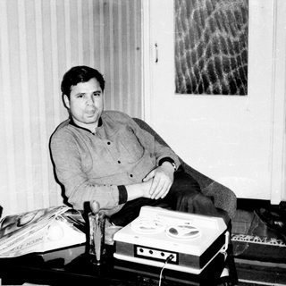 Man seated with vintage reel-to-reel recording equipment.