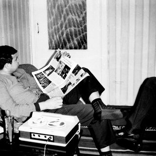 Man reading newspaper in recliner with reel-to-reel tape recorder.