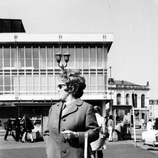 Woman in 1960s coat standing near bus station.