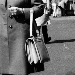 Woman in 1960s coat standing near bus station.