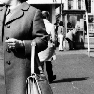 Woman in 1960s coat standing near bus station.