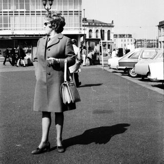 Woman in 1960s coat standing near bus station.