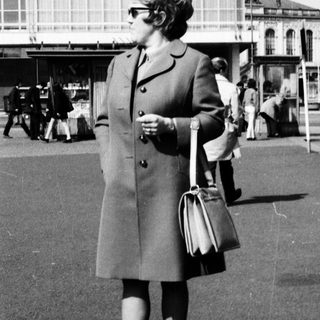 Woman in 1960s coat standing near bus station.