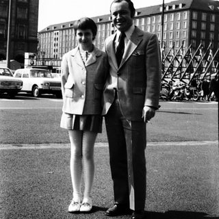 Two people posing together in formal attire, 1950s-60s urban setting.